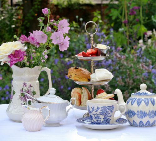 Photograph of a table set for afternoon tea. The table is covered with a white table cloth and there is a three-tiered cake stand containing a sandwich, a scone, a meringue, a cake and some strawberries and cream. There is a blue and white teapot and cup and saucer and a white china milk jug and sugar bowl. To the left there is a jug containing an arrangement of flowers. The table is standing in an English country garden. Flowers can be seen in the background.