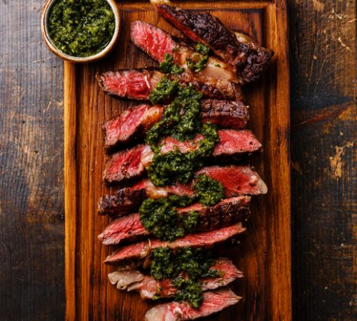 Sliced medium rare grilled beef barbecue Ribeye steak with chimichurri sauce on cutting board on dark background